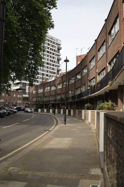 The image shows a curved residential street lined with multi-storey brick apartment buildings with black metal balconies. The pavement features a wide sidewalk with a tactile paving strip near the cur