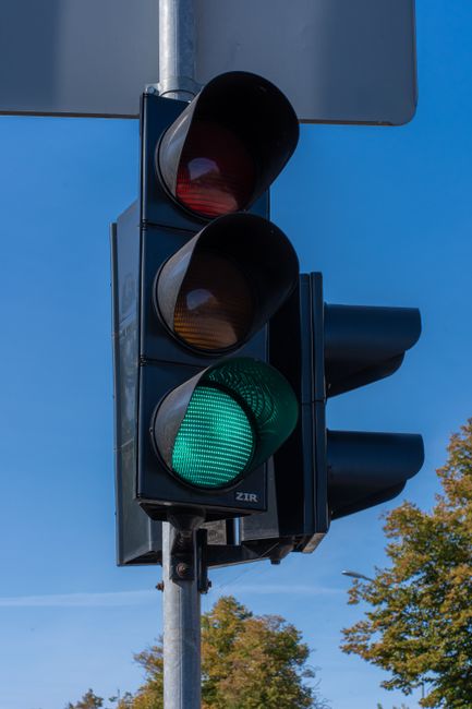 A close-up image of a pedestrian traffic light installed on a metal pole, displaying a green signal indicating it is safe to cross. The traffic light has three circular lights stacked vertically, with